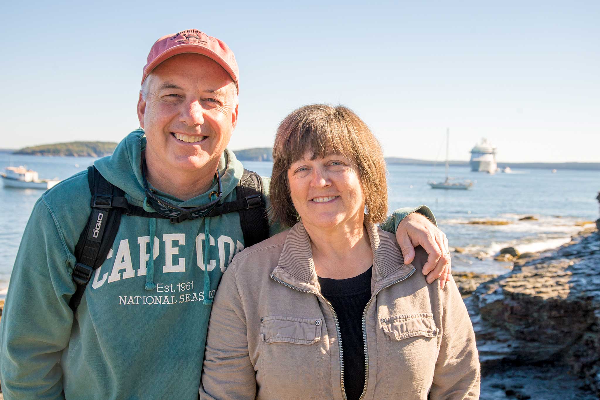 A couple in their early 60s smile for the camera with the man’s arm around his wife’s shoulder; a sunny, blue bay with small green hills and various boats are in the background.