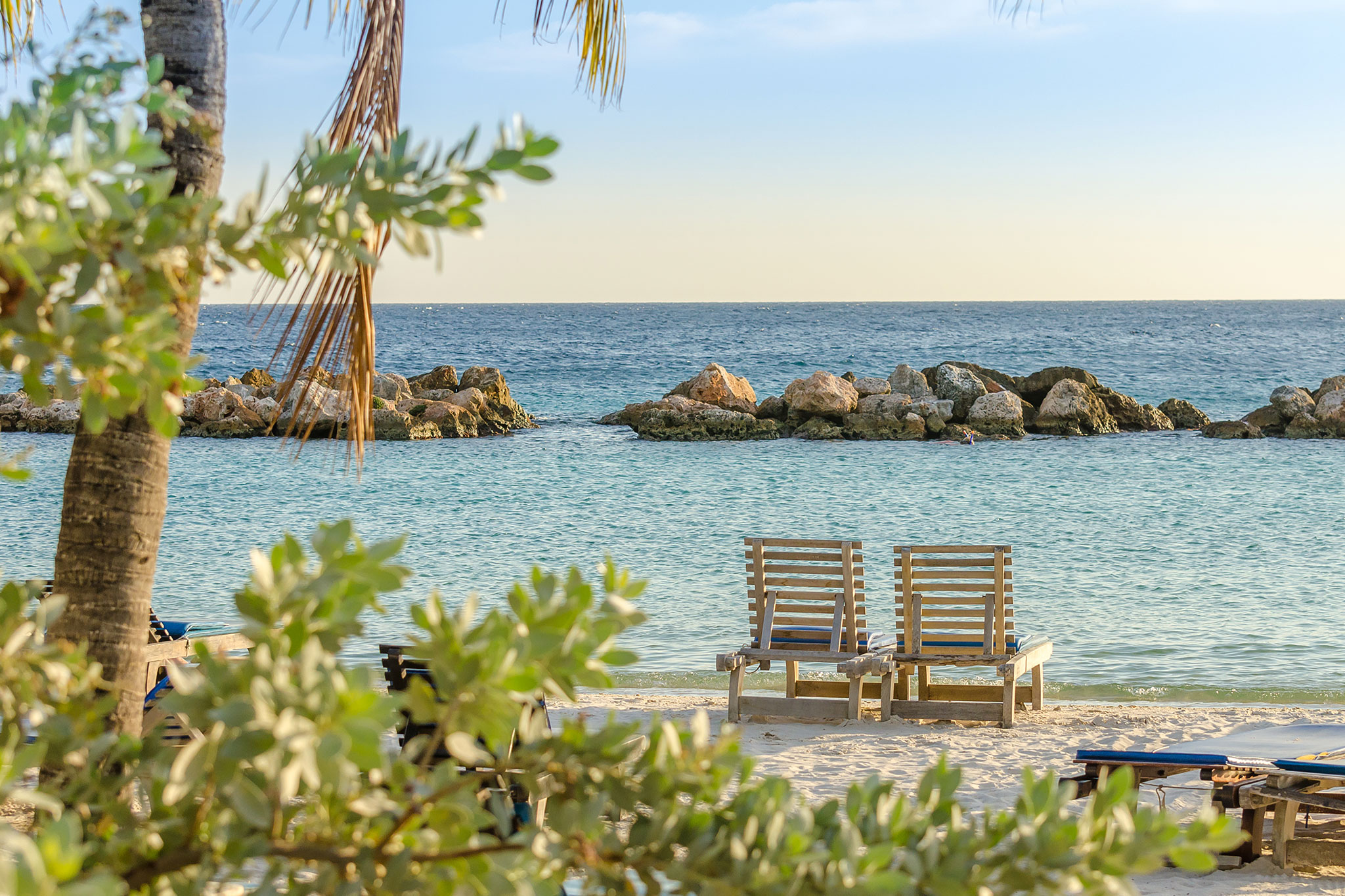 A peaceful seaside view featuring a relaxing setup of two beach chairs on sandy shores with clear turquoise waters and rocks, framed by tropical plants and palm trees.