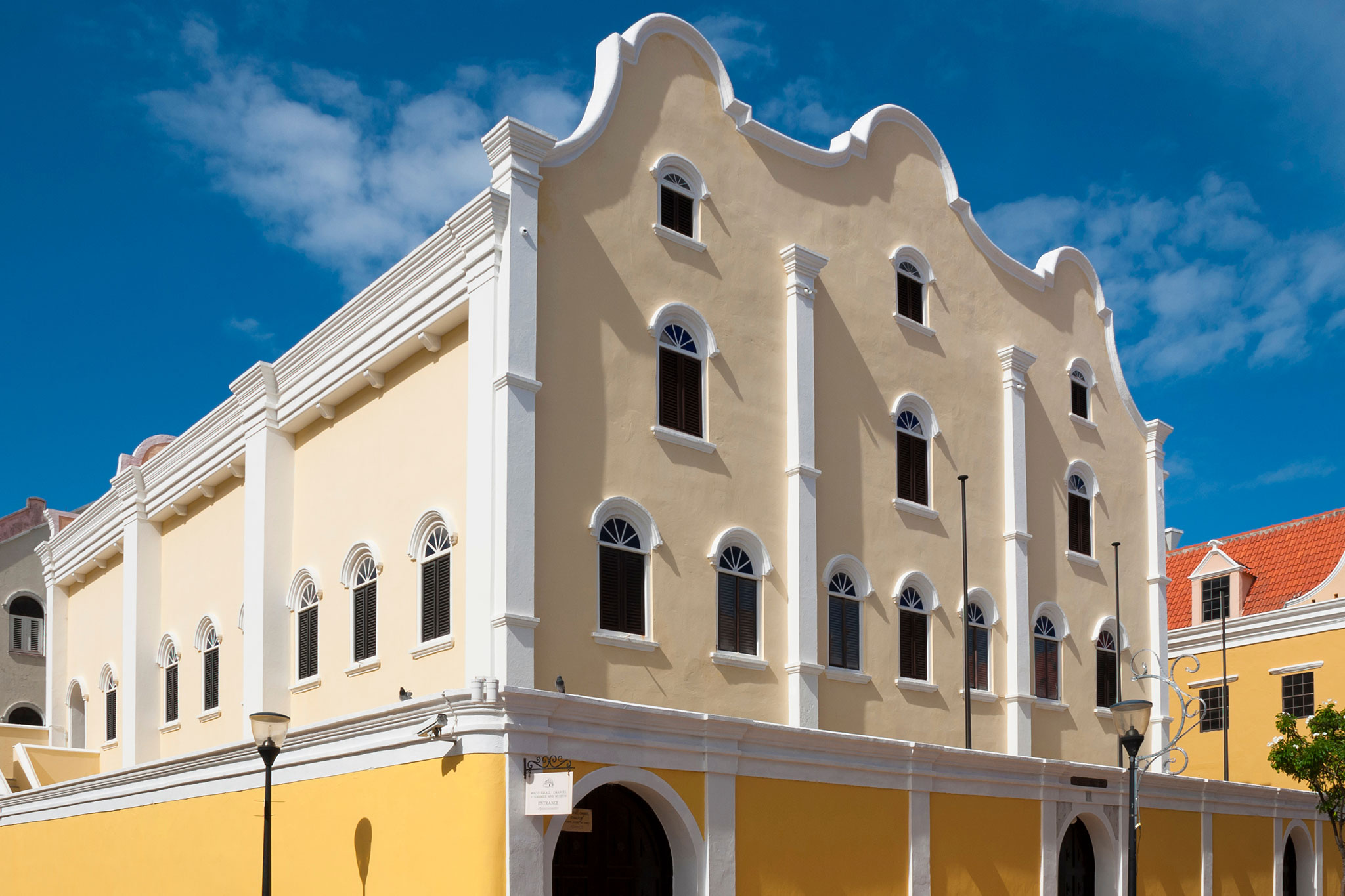 Elegant yellow-and-white synagogue with ornate arched windows and baroque architectural details, set against a blue sky.