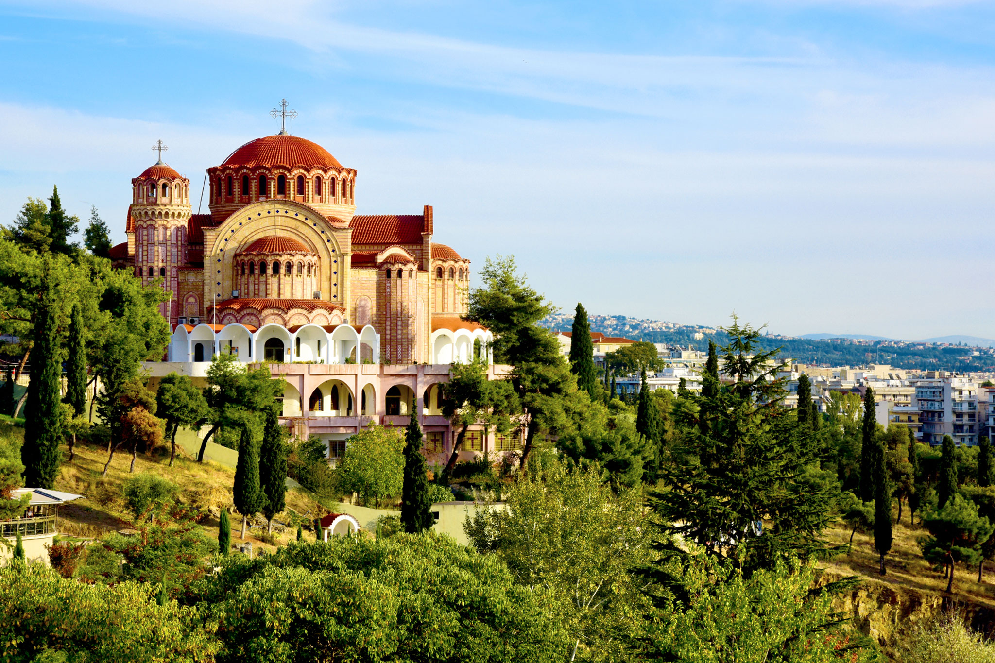 Scenic view of Saint Paul’s Orthodox Cathedral in Thessaloniki, Greece, showing the church’s distinctive ornate red domes and Byzantine architecture, nestled among lush greenery with the cityscape in the background.