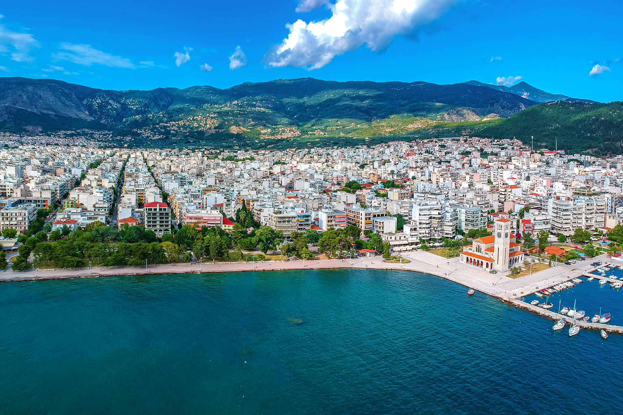 An aerial view of Volos, Greece, showing the expansive cityscape nestled between the sea and rolling hills. It features a prominent red-roofed church near the coastline.