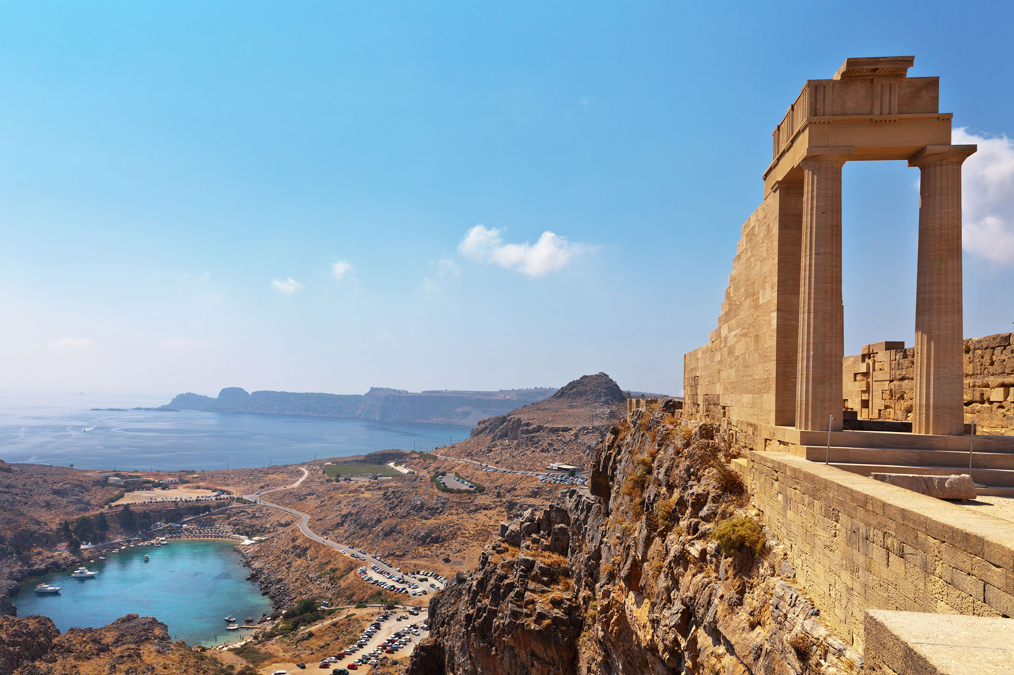 Ancient stone columns of the Acropolis of Lindos on the island of Rhodes, Greece, overlooking the serene St. Paul's Bay with a clear view of the rugged coastline and the Aegean Sea.