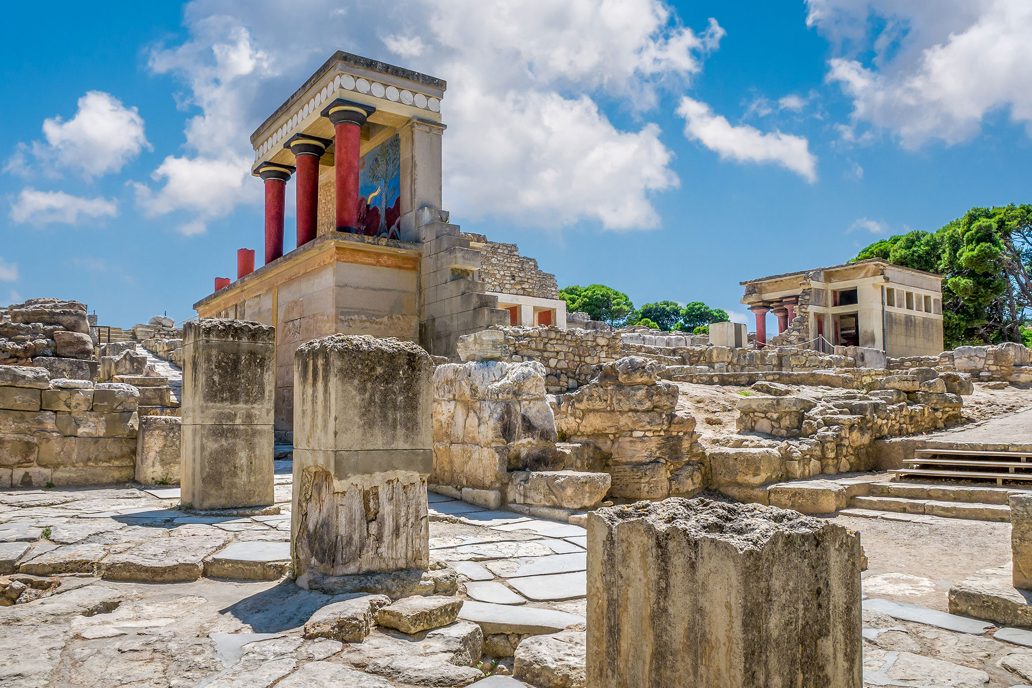 Sunny day at Knossos Palace, Crete, highlighting the restored architectural details with colorful columns and frescoes, surrounded by the ruins of the ancient Minoan civilization.