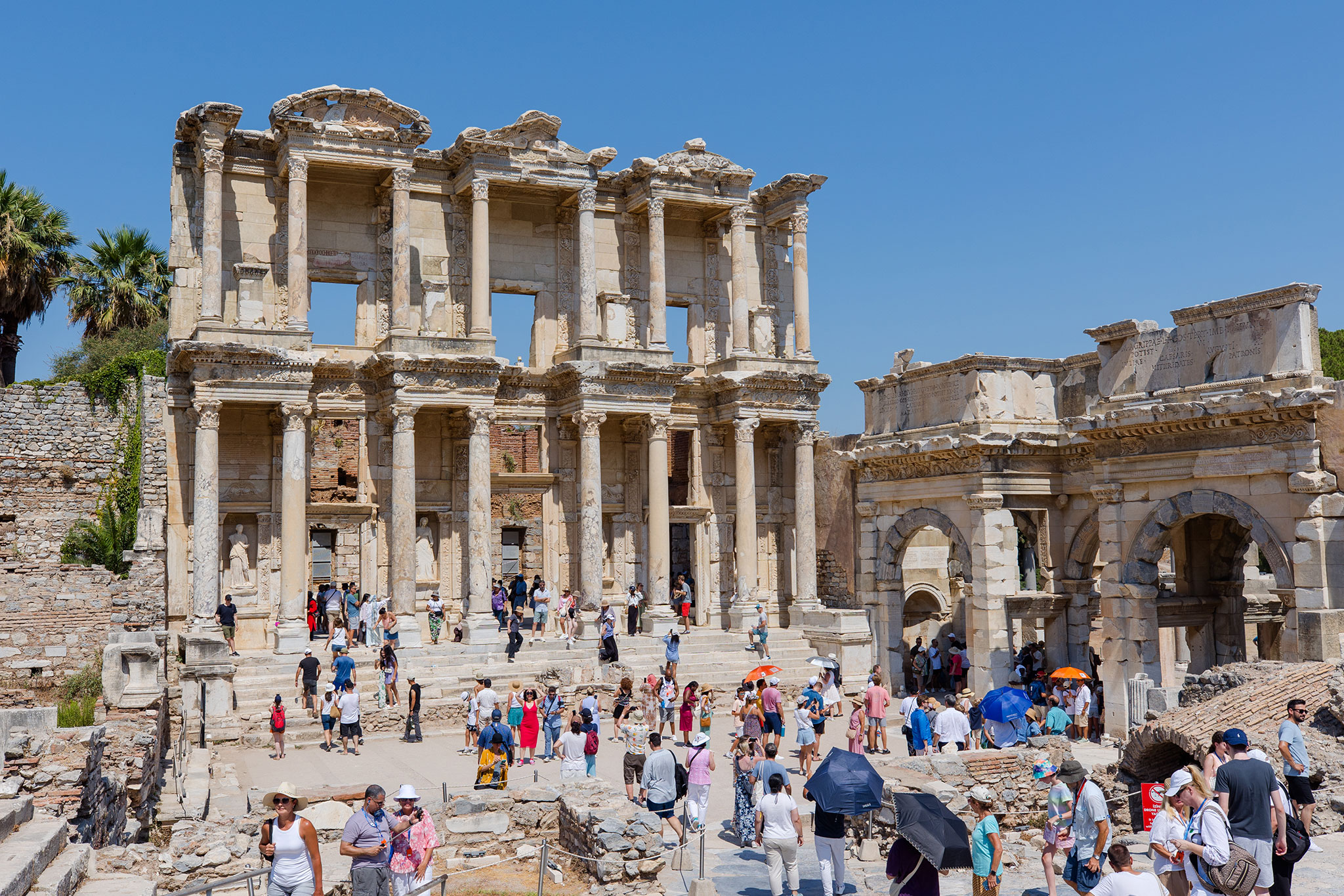 Busy scene at the Library of Celsus in Ephesus, with travelers walking among the towering columns and archaeological remnants.