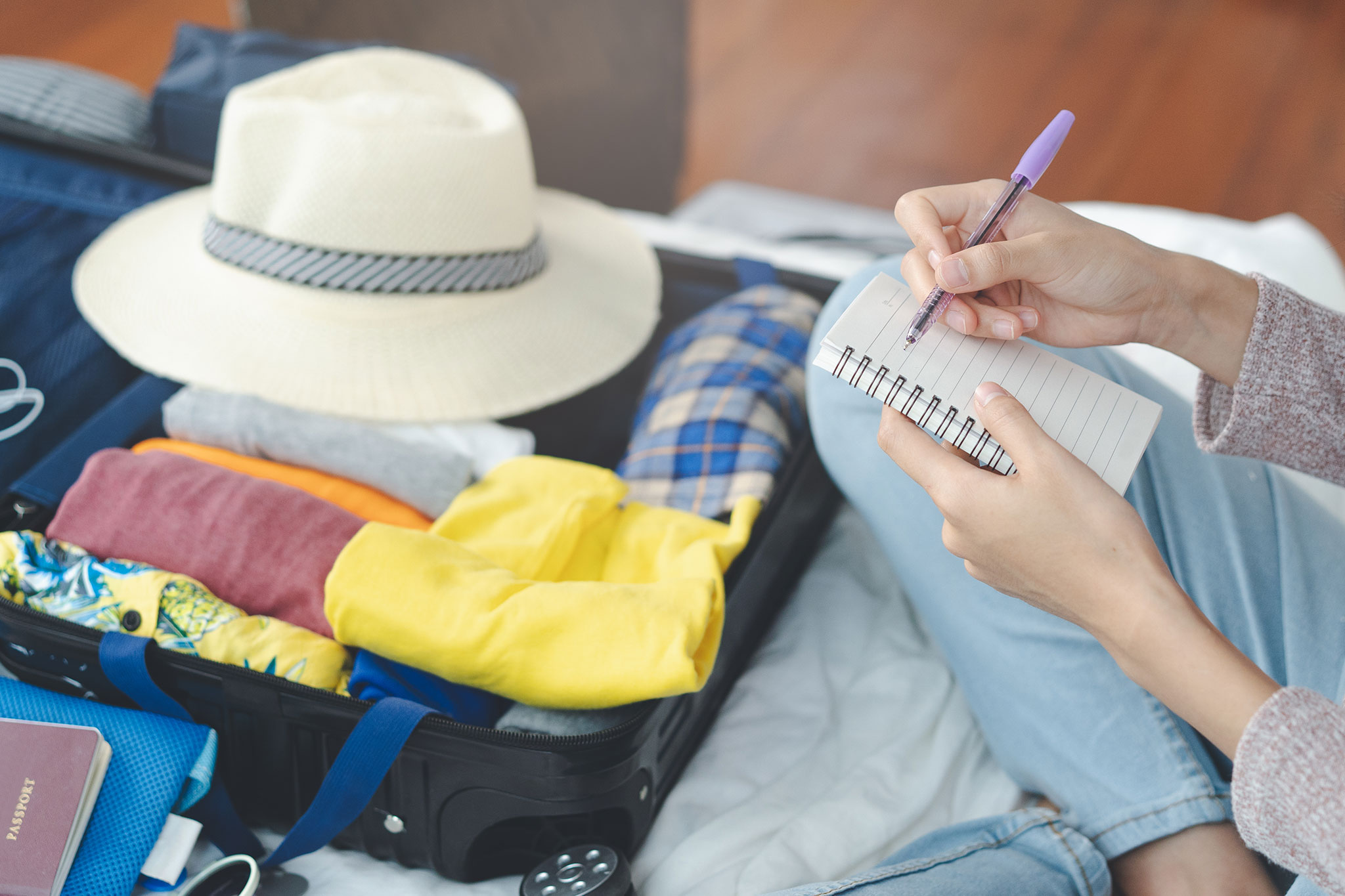 A young woman sits on a bed and uses a list to check accessories and clothing in her luggage before traveling.