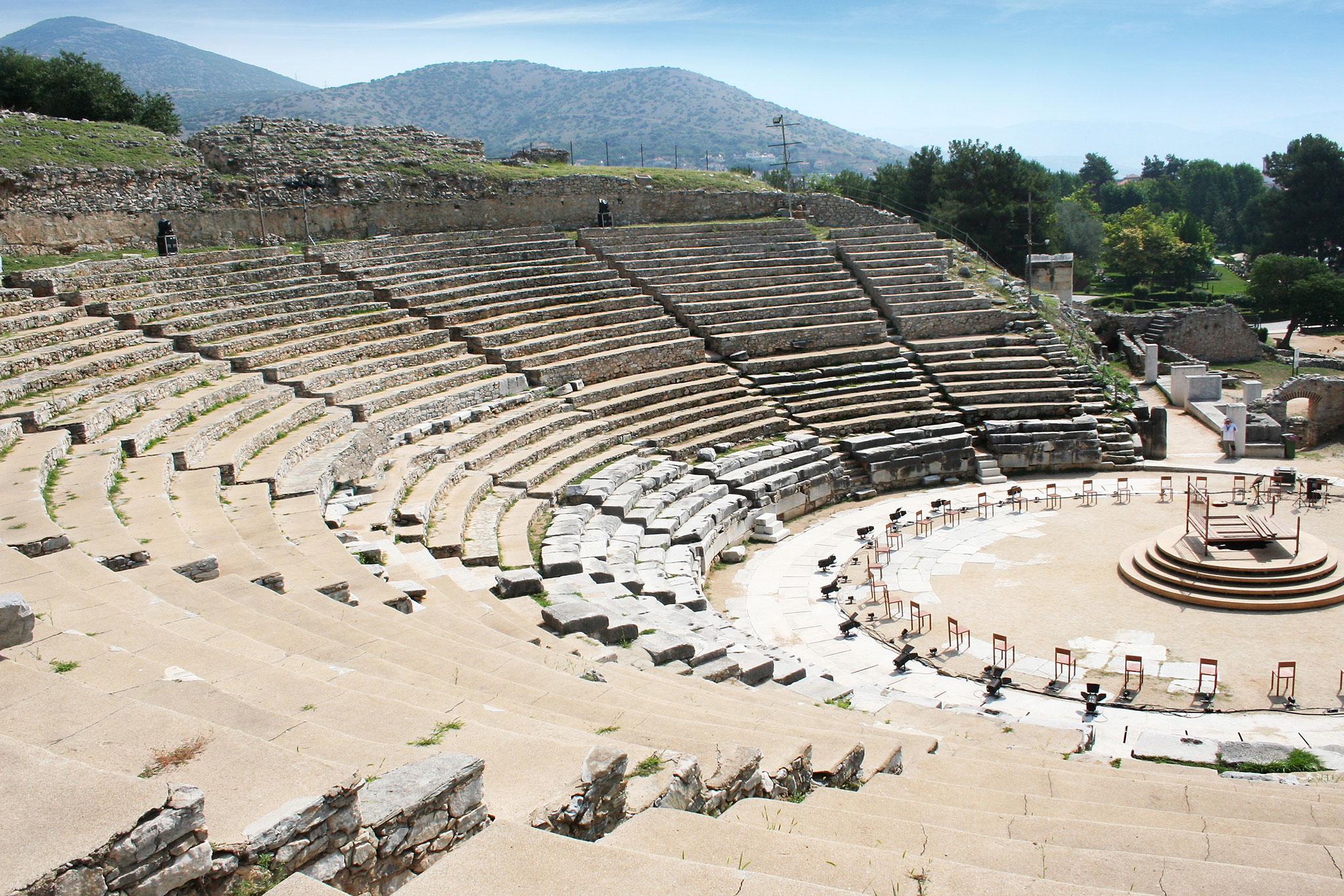 Ancient amphitheater with tiered seating and a central stage, nestled in a natural landscape, with mountains in the background on a sunny day.