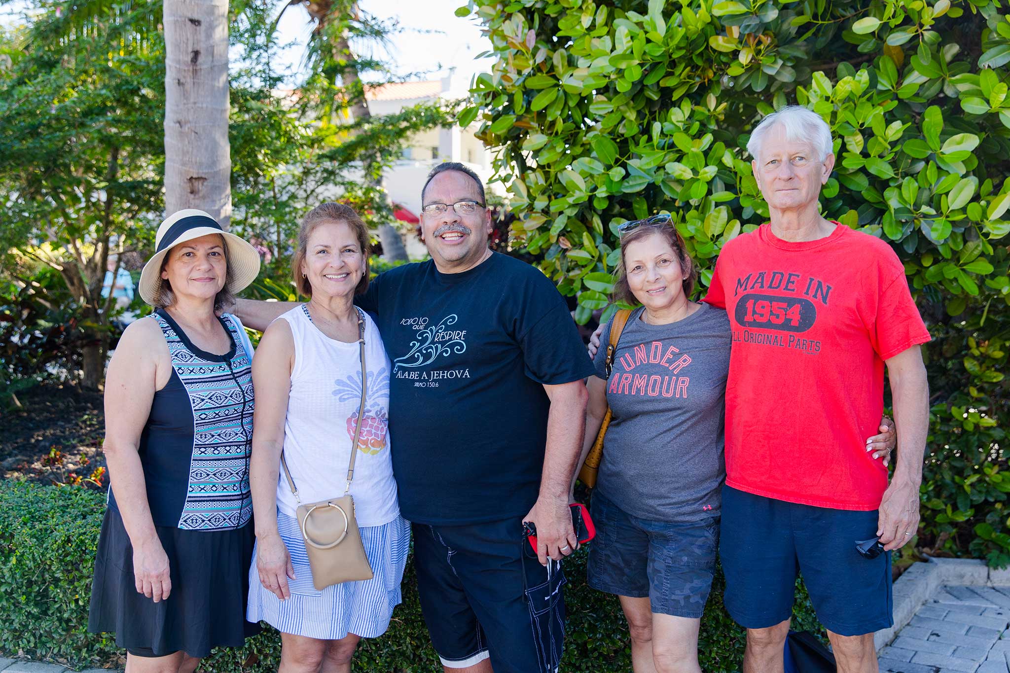 Three women and two men posing for a photo under a tree in a tropical garden.