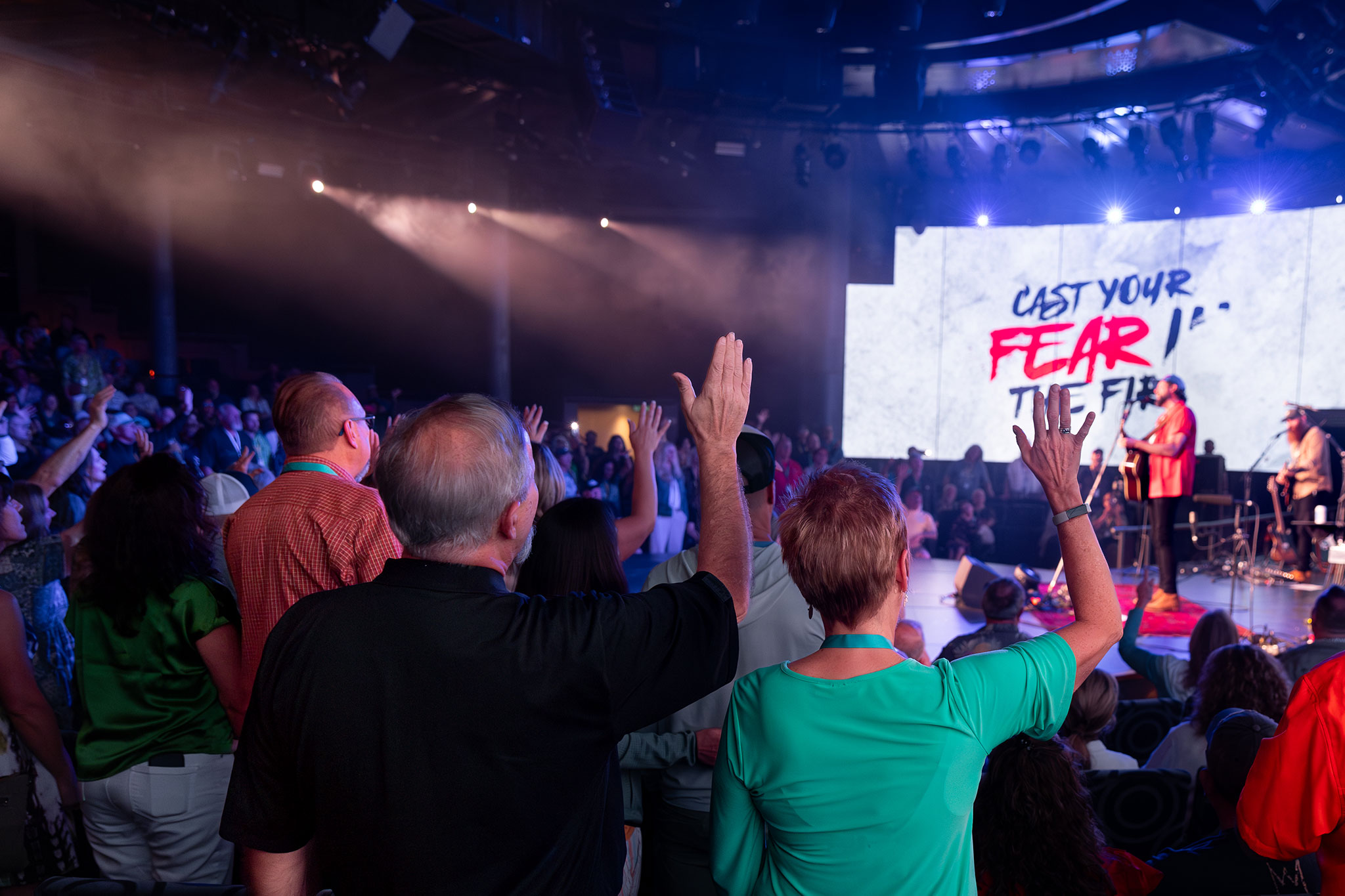 Audience members raise their hands in a vibrant indoor concert setting, illuminated by stage lights and smoke, with a backdrop displaying song lyrics.