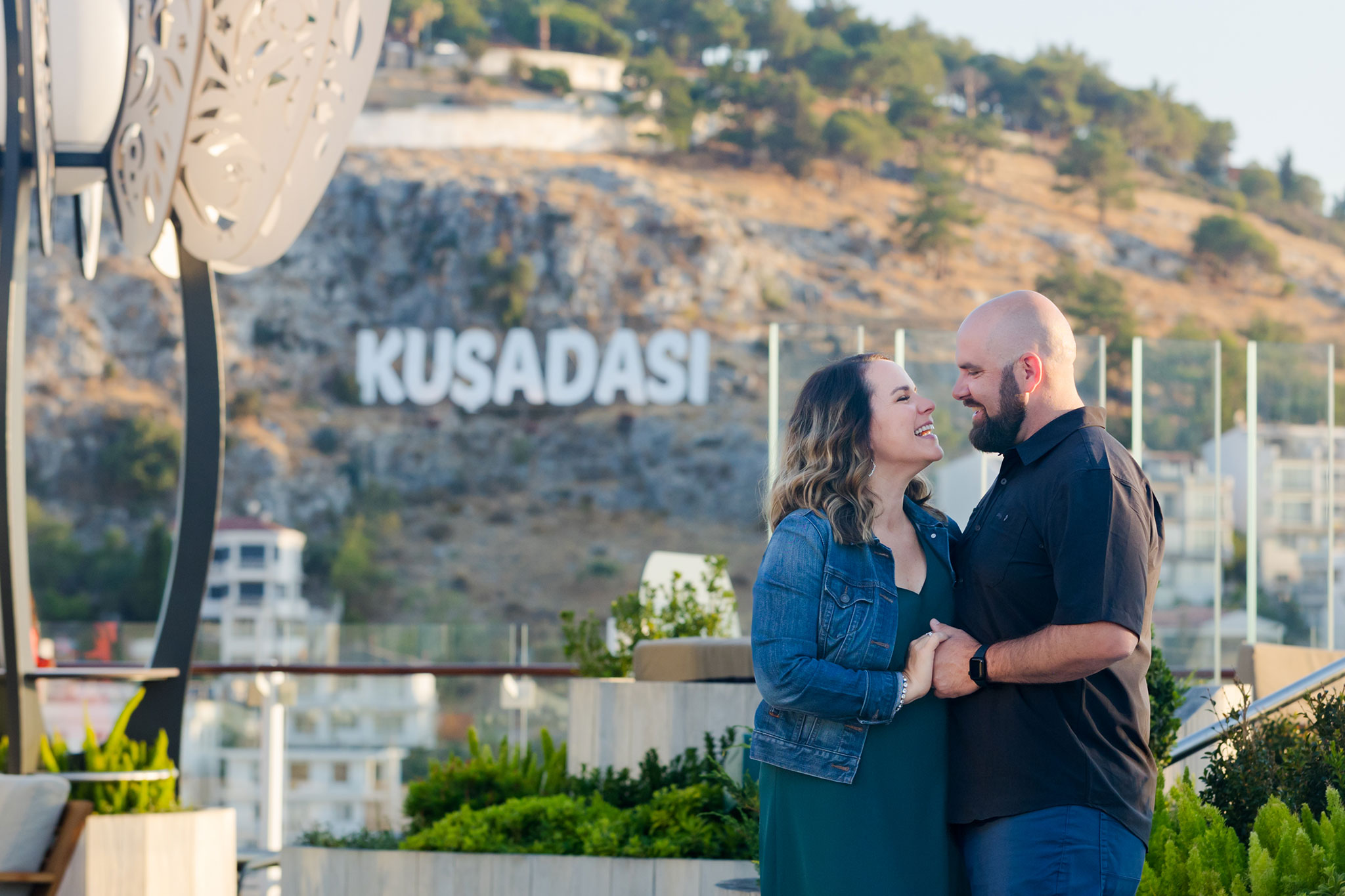 A couple smiling at each other, embracing on a terrace with the Kusadasi sign on a hill in the background.