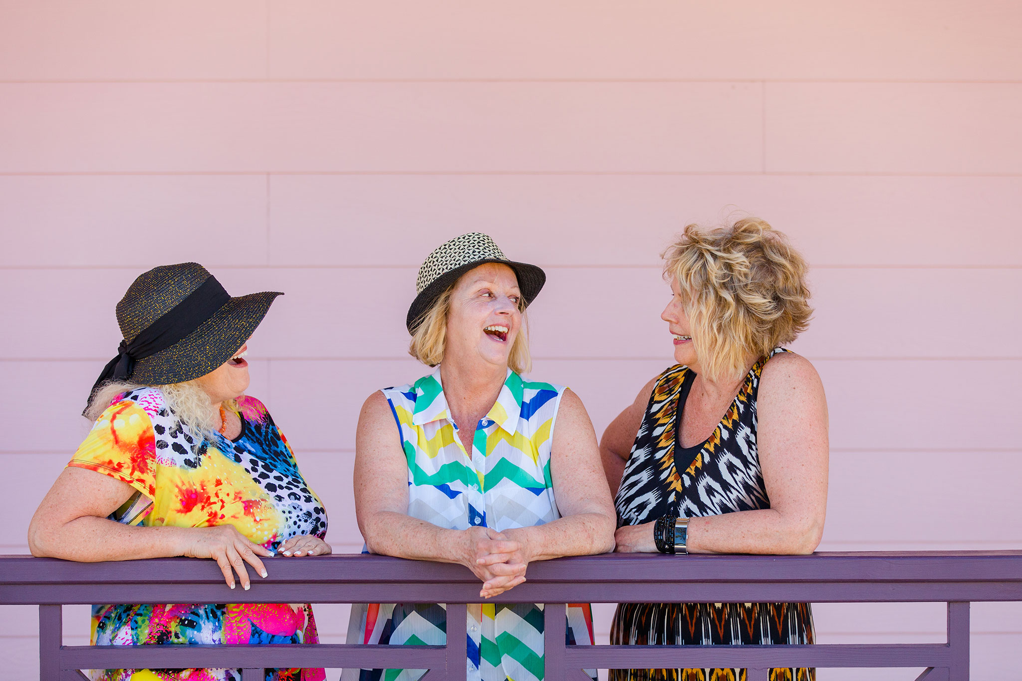 Three senior women laugh and chat together, leaning on a railing, wearing colorful summer outfits and stylish hats against a pale pink wall.