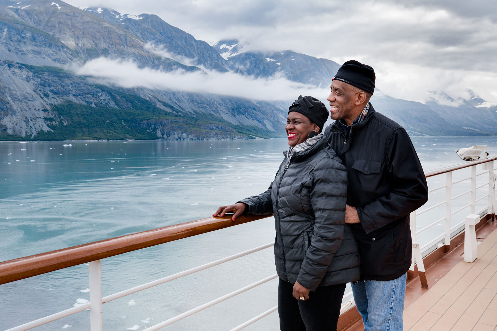 An elderly couple joyfully leaning on the railing of a cruise ship with a stunning view of a glacier and mountains in the background.