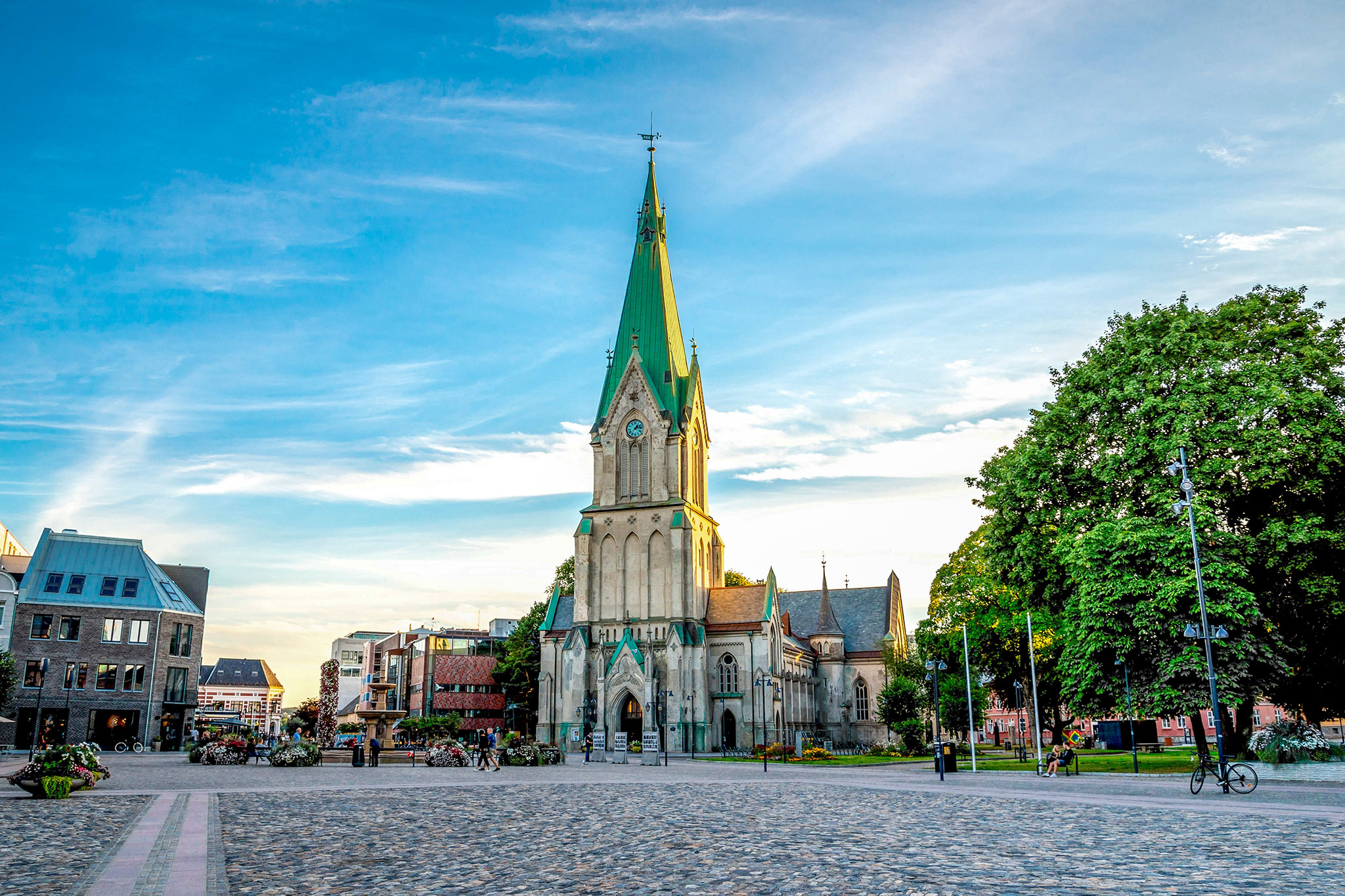 Neo-Gothic Kristiansand Cathedral with its stone facade and soaring green spire stands in the heart of the city, framed by leafy trees and modern buildings.