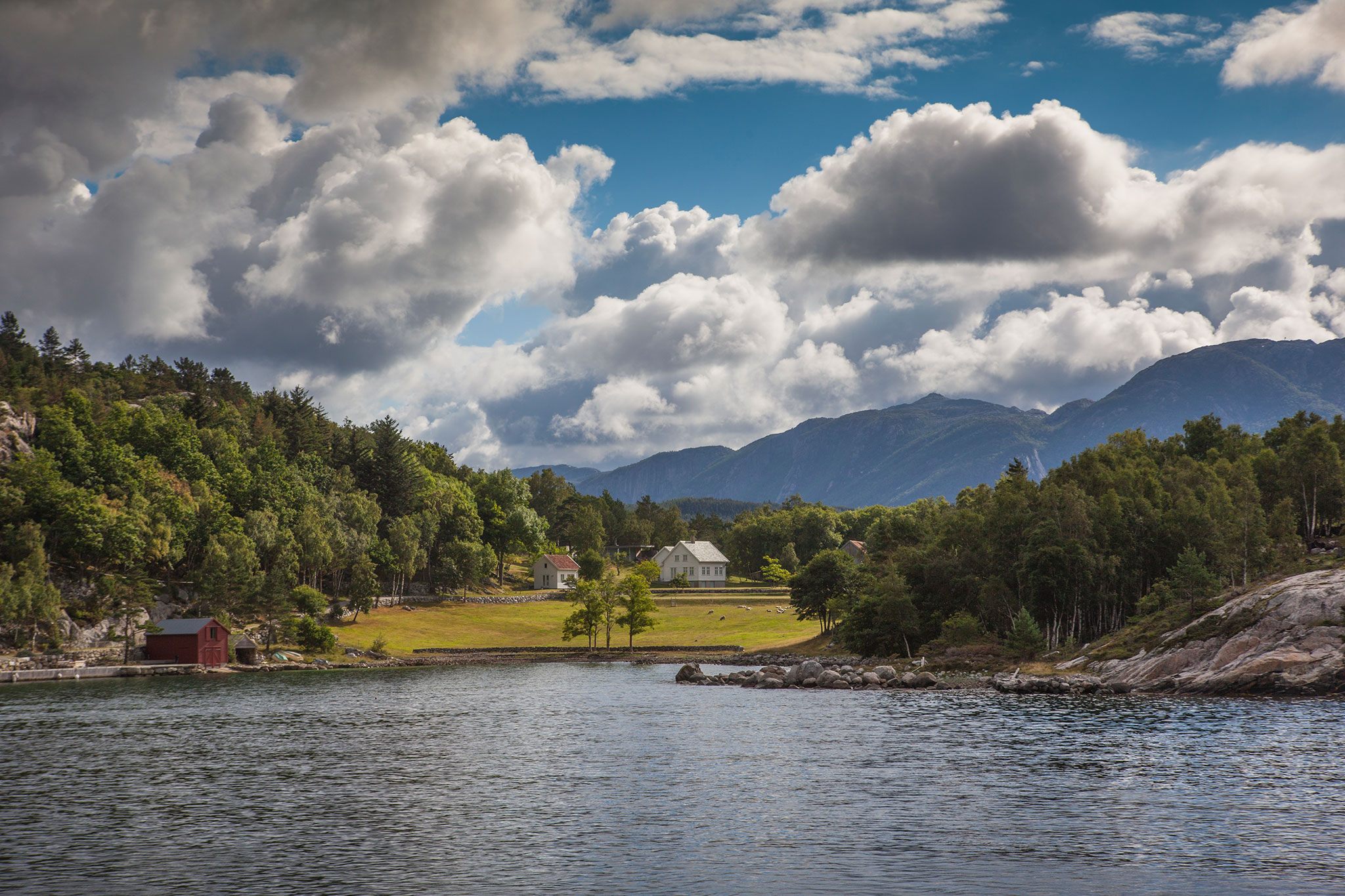 Scenic Norwegian fjord with a white farmhouse and red boathouse set among grassy fields, green forests, and mountains under dramatic clouds.