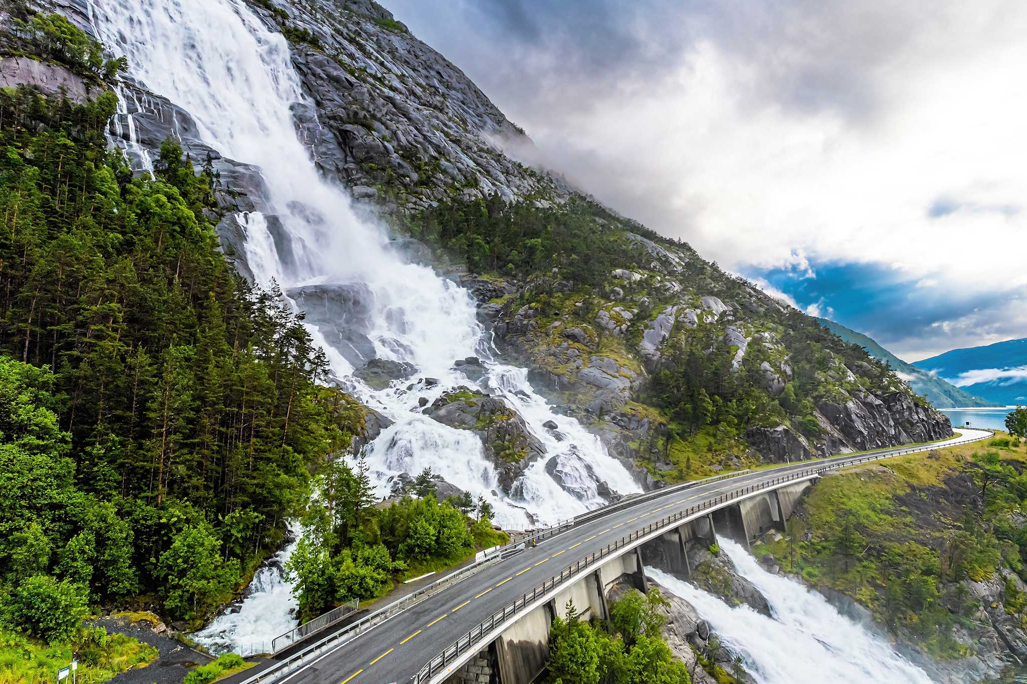 Majestic waterfall cascades down a rocky mountainside beside a winding highway, surrounded by lush green forest and fjord views.