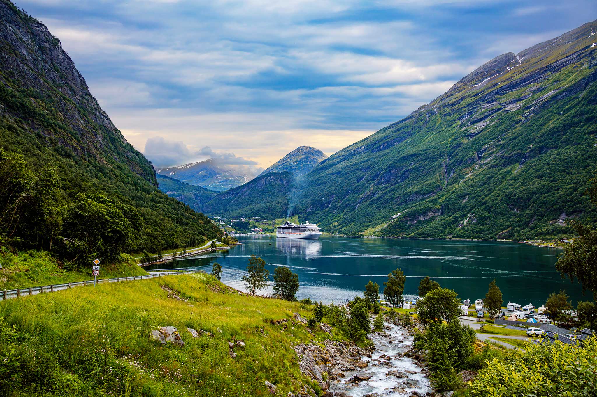 Cruise ship sailing through a narrow fjord surrounded by steep, green mountains, with calm, reflective water, a small riverside village and dramatic clouds overhead.