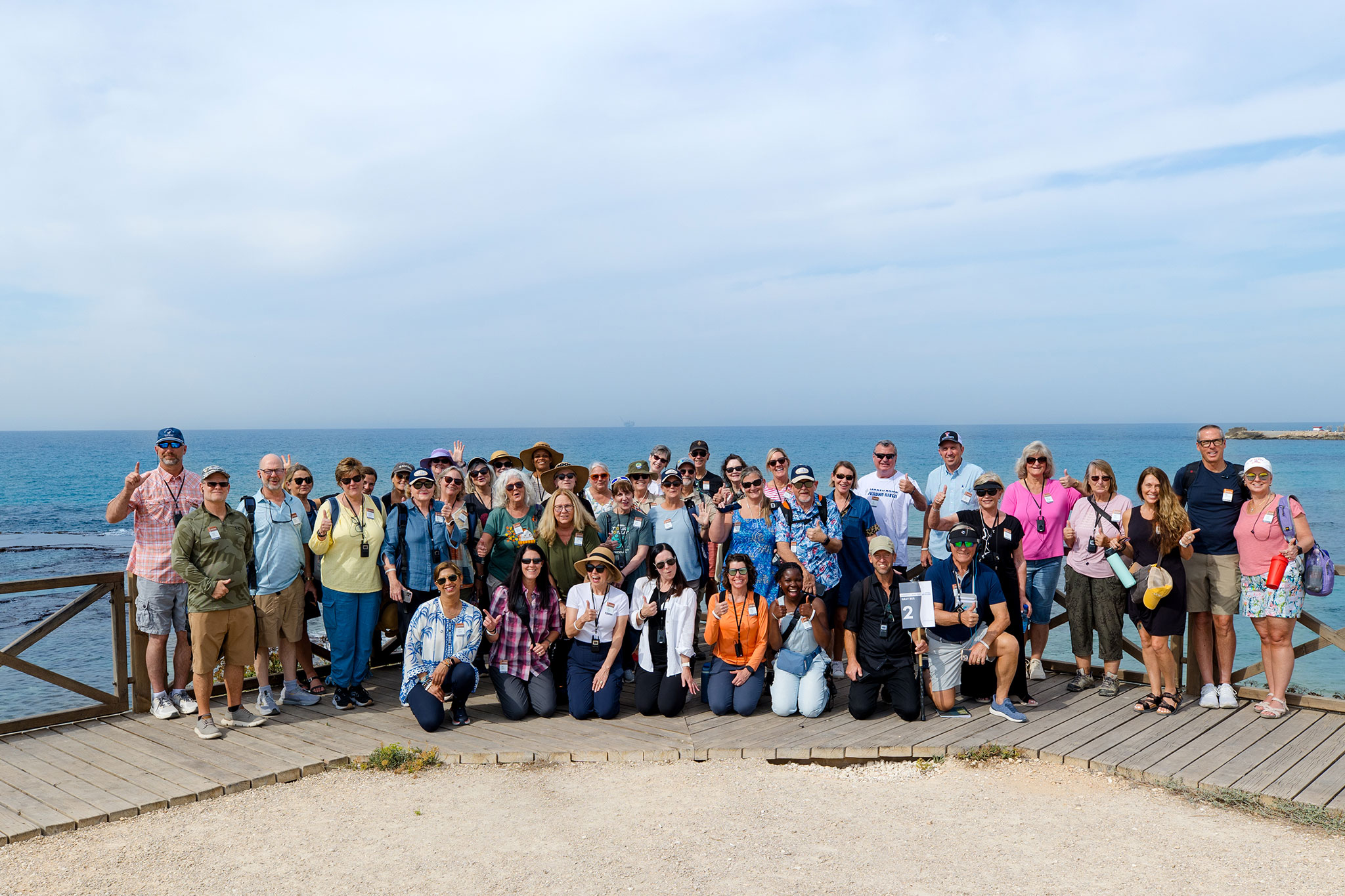 A large group of travelers poses together on a wooden boardwalk at Caesarea Maritima, smiling at the camera, with the Mediterranean Sea and a clear blue sky behind them.