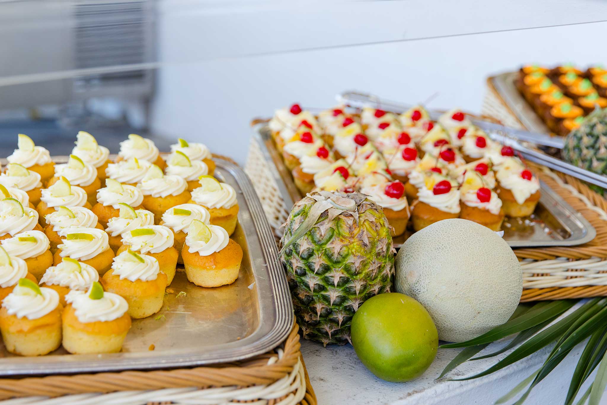 Trays of bite-sized frosted desserts topped with citrus and cherries sit on a buffet table alongside fresh tropical fruit, including pineapple, melon, and lime.