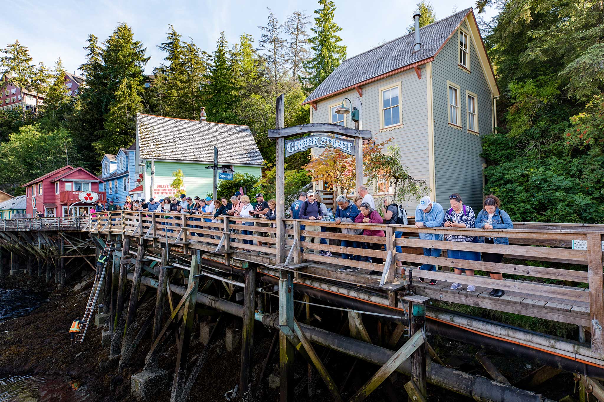 Visitors lining a wooden boardwalk on Creek Street in Ketchikan, Alaska, look down at the water below, with colorful historic houses on stilts, a sign that reads “Creek Street,” and a lush forested hillside in the background.