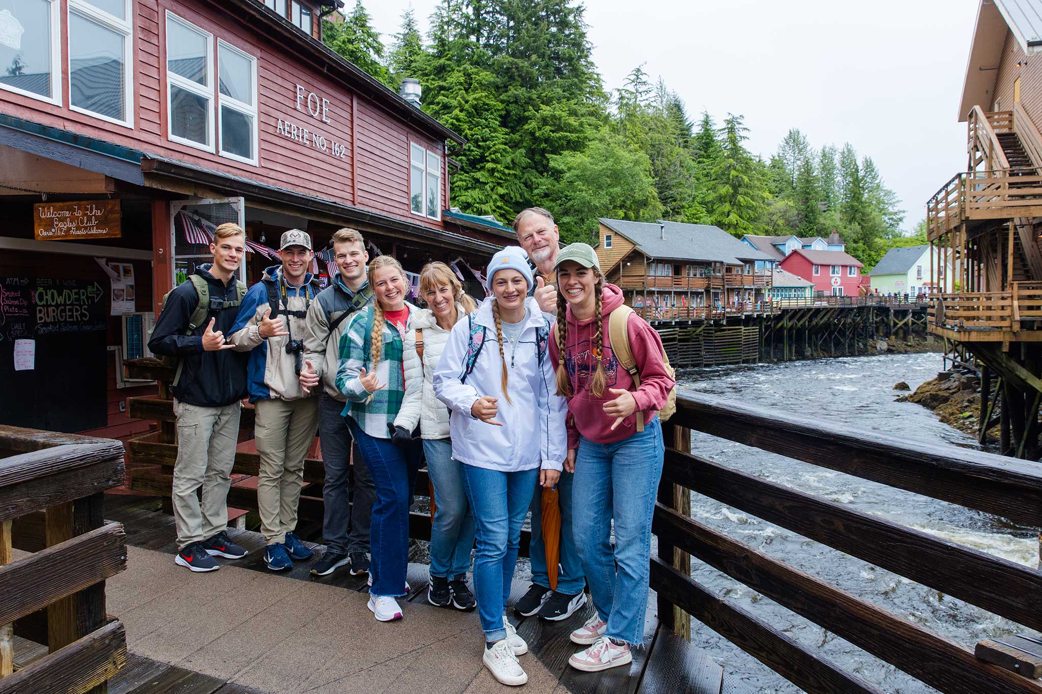 A group of smiling travelers poses on a wooden boardwalk in Ketchikan, beside Creek Street’s colorful buildings on stilts above rushing water, with forested hills and historic storefronts in the background.