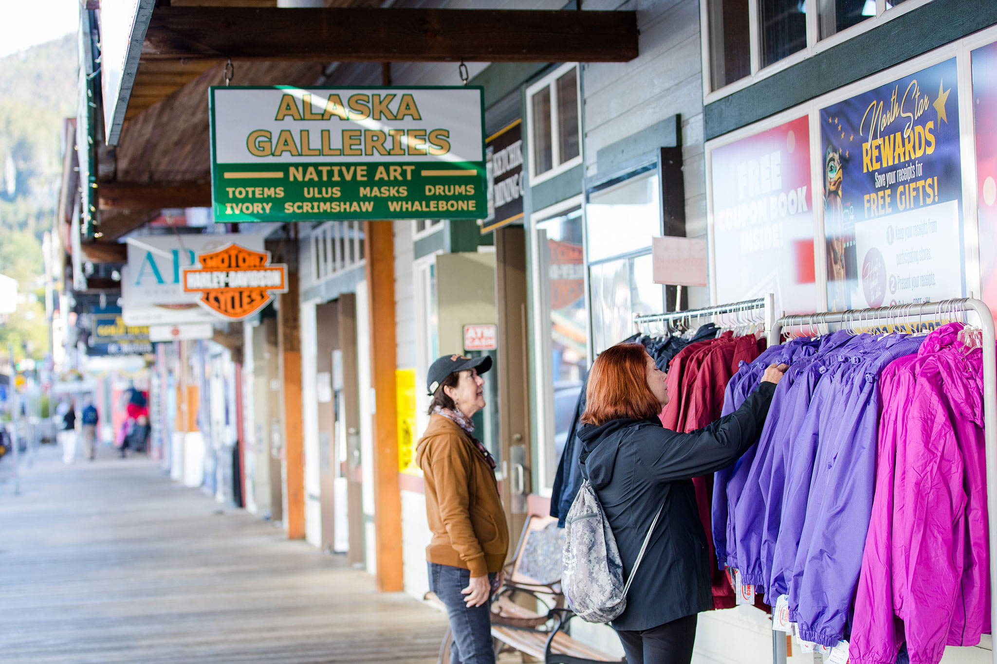 Two travelers browse colorful jackets outside shops along a wooden boardwalk in Ketchikan, Alaska, with storefront signs including “Alaska Galleries Native Art” and mountain scenery in the background.
