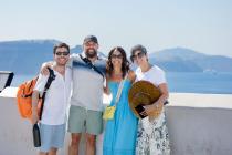 A group of four tourists smiling and posing for a photo in Oia Village, Santorini, with the stunning blue waters of the Aegean Sea and the caldera in the background.