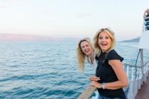 Two smiling women laugh and lean on the railing of a boat with scenic ocean views behind them.
