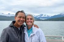 Smiling couple poses together on a ship’s deck with snowy mountains and water in the background.
