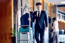 A hotel staff member in a suit smiles while pushing a luggage cart loaded with suitcases through a modern hotel lobby.