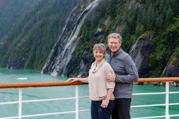 Smiling couple on a cruise deck with emerald fjord waters, rocky cliffs, and waterfalls in the background.