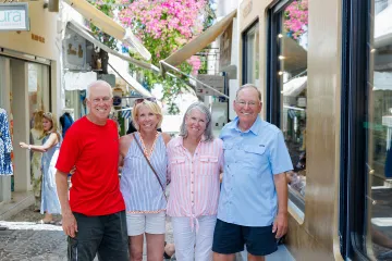 Four smiling travelers pose together on a sunny cobblestone street lined with shops and flowers.
