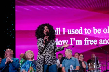 Lynda Randle sings passionately onstage with a microphone, backed by seated performers and a bright pink screen displaying song lyrics.