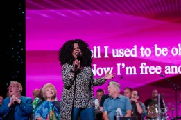 Lynda Randle sings passionately onstage with a microphone, backed by seated performers and a bright pink screen displaying song lyrics.