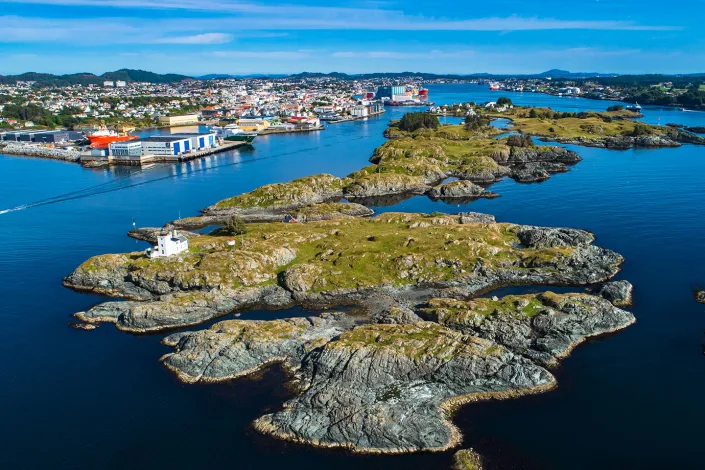 Aerial view of small rocky islands with a white lighthouse near Haugesund, Norway, surrounded by calm blue water and distant cityscape