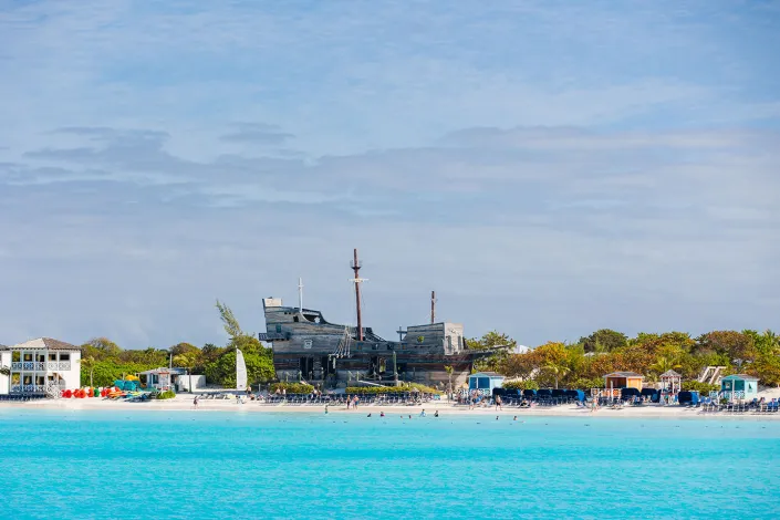 A bright turquoise shoreline with calm ocean water in the foreground, beach chairs and small colorful buildings along the sand, and a large wooden pirate ship structure set among palm trees under a blue, lightly clouded sky.