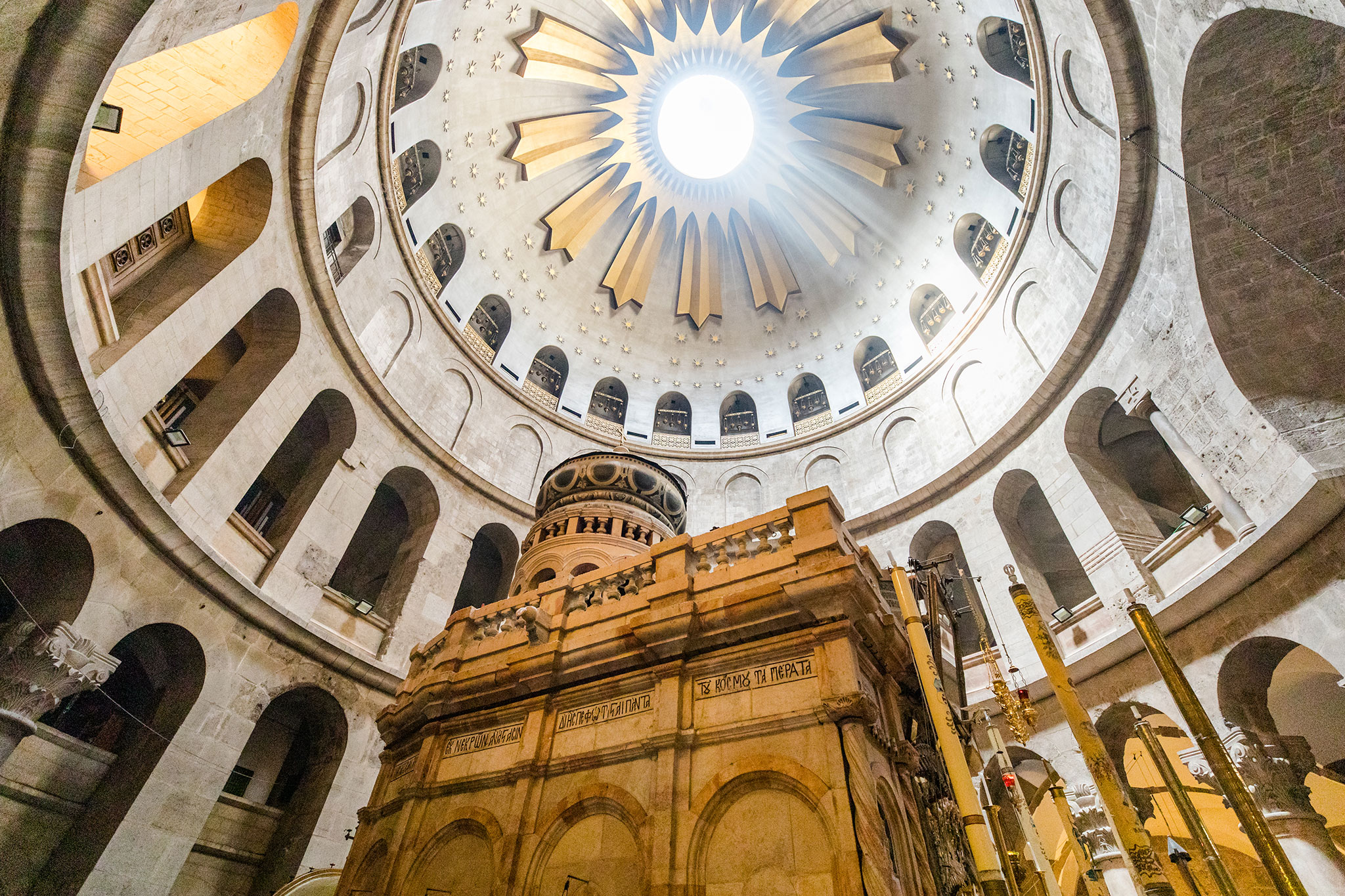 Interior view of the Church of the Holy Sepulchre rotunda, looking upward toward a bright circular oculus that floods the domed ceiling with light. Below, the ornate stone Edicule marking the tomb stands at the center, surrounded by tall columns, arched galleries, and warm-toned lamps.