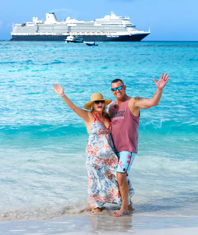 A couple smiling and waving on the beach with a large cruise ship in the background