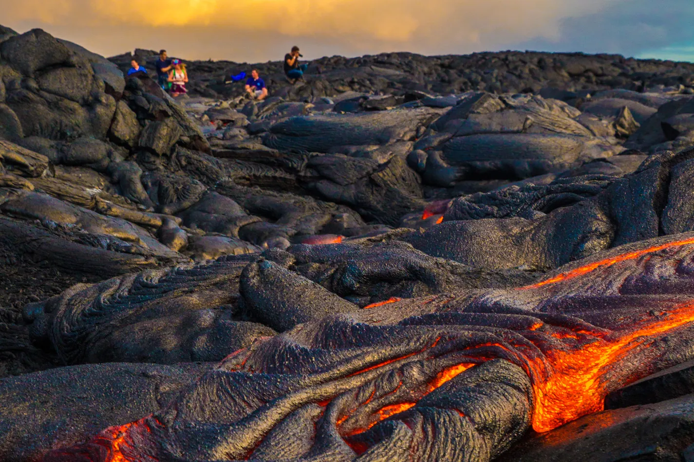 Glowing orange lava flows between hardened black volcanic rock while a group of people observe from a safe distance under a cloudy, golden sky.