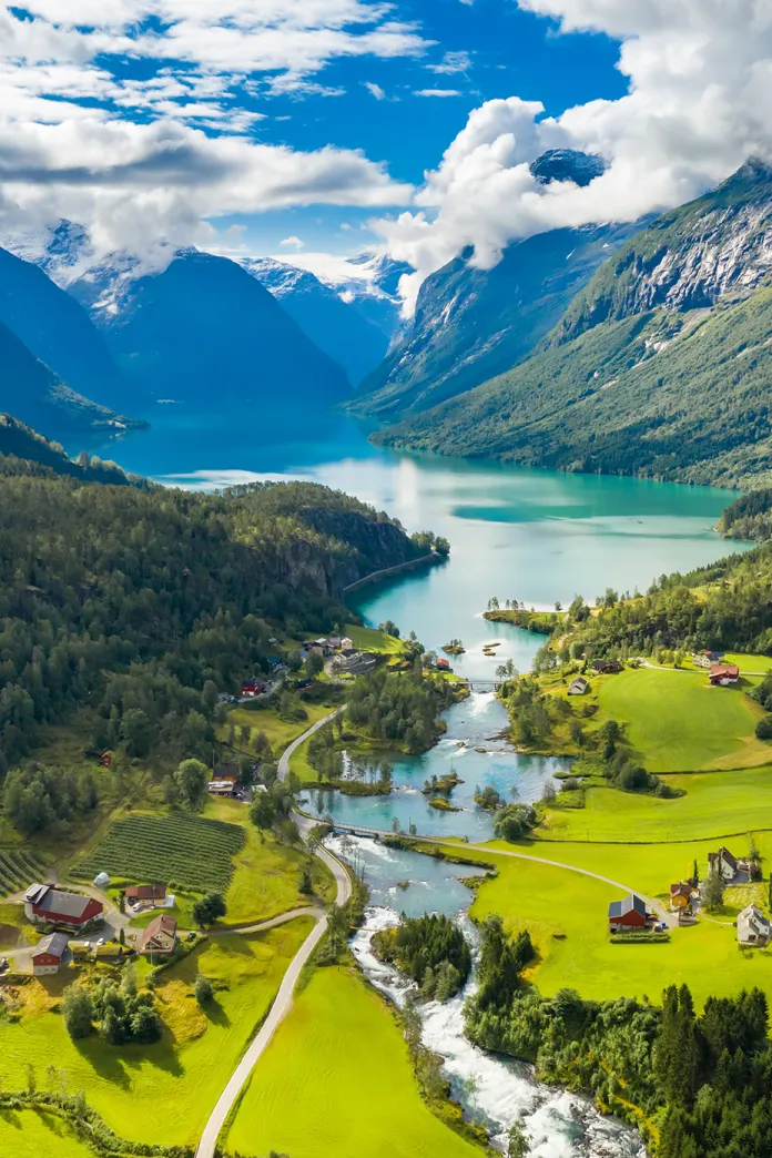 Scenic aerial view of a turquoise fjord surrounded by lush green valleys, winding roads, and snow-capped mountains in Norway under a bright blue sky with scattered clouds.