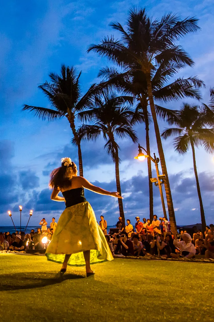 A dancer in a yellow skirt performs a traditional hula at dusk, illuminated by warm lights, with palm trees silhouetted against a deep blue sky and a seated crowd watching in the background.