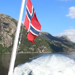 The Norwegian flag blowing in the wind with a large body of water and a mountain in the background