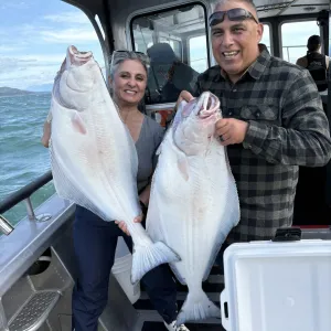 A smiling man and woman stand on a fishing boat holding two large halibut.